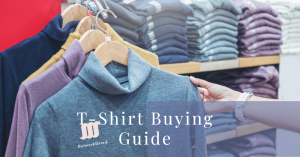 Hand holding a grey t-shirt on a hanger with assorted folded t-shirts in the background.
