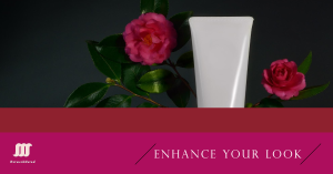 A white cosmetic tube placed between two vibrant pink flowers with green leaves, set against a dark background, accompanied by the text "Enhance Your Look."