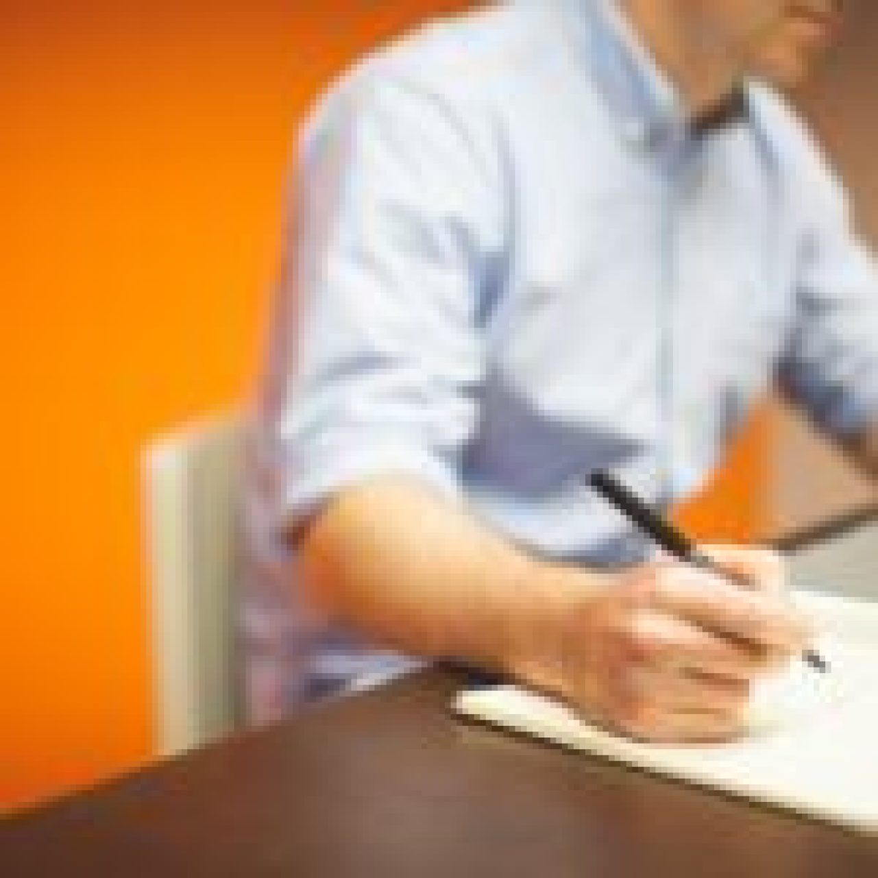 A person in a blue shirt is seated at a desk, writing in a notebook with the left hand while using a laptop with the right hand. The background features an orange wall, providing a warm contrast.