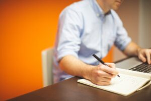 A person in a blue shirt is seated at a desk, writing in a notebook with the left hand while using a laptop with the right hand. The background features an orange wall, providing a warm contrast.