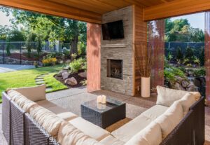 Modern outdoor patio with wicker furniture and stone fireplace under wooden pergola surrounded by green garden.