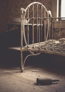 An old, rusty metal bed frame in a dimly lit, abandoned room, surrounded by dust and debris.