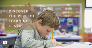 Child writing at a desk in a classroom with colorful decorations in the background.