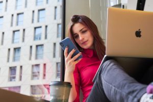 Person working remotely with a laptop and smartphone, sitting comfortably by a window with a cityscape view.