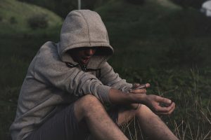 An individual sitting on the ground outdoors, wearing a hoodie with their face obscured, holding what appears to be a syringe, illustrating the struggles associated with drug use and the importance of understanding rehab stigmas.