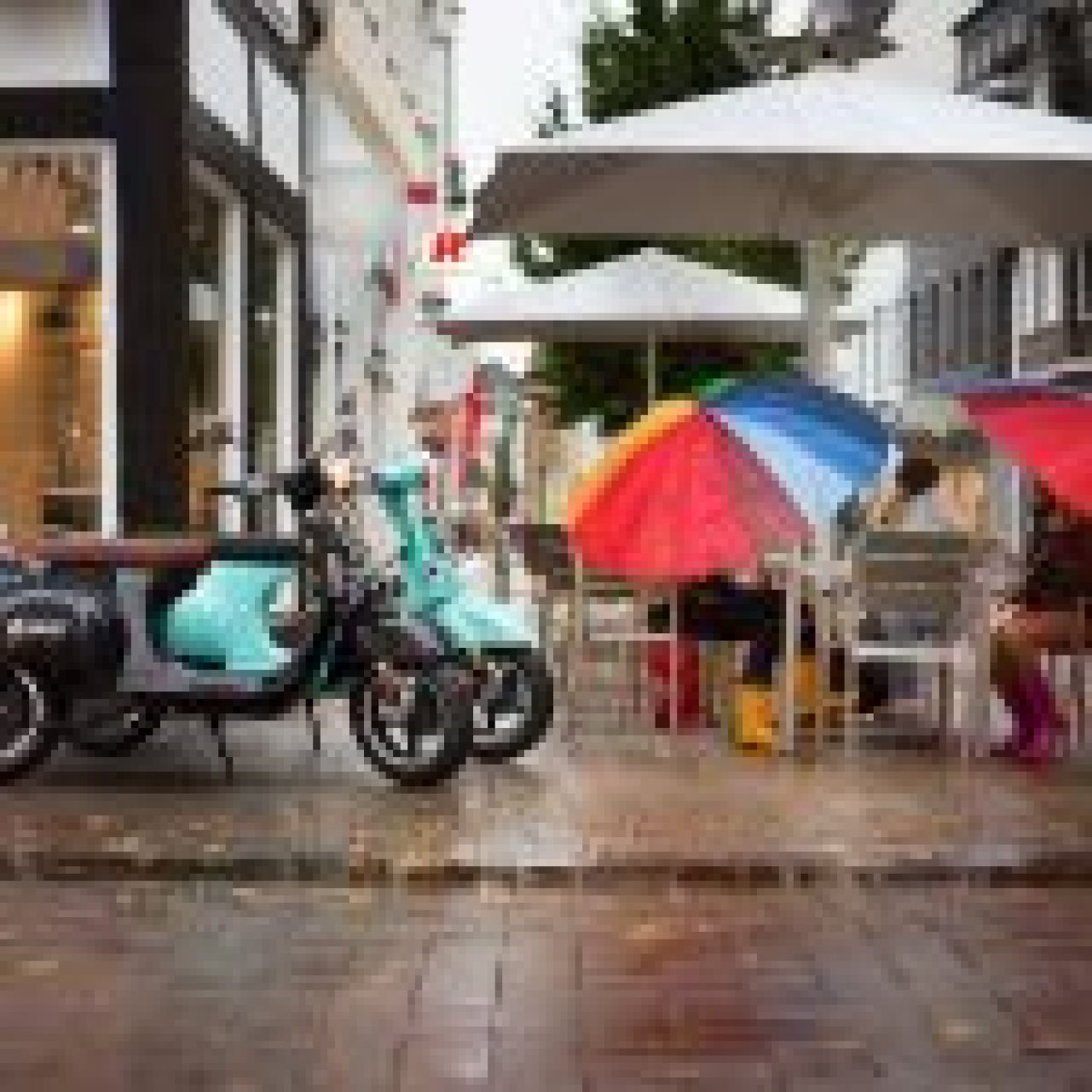 A teal electric scooter parked on a wet city street next to a sidewalk café with colorful open umbrellas.