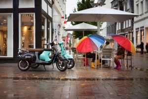 A teal electric scooter parked on a wet city street next to a sidewalk café with colorful open umbrellas.