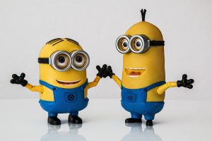 Two Minion figurines standing side by side on a reflective surface against a white background. One Minion has two eyes and is raising its arms, while the other has one eye and an open mouth.