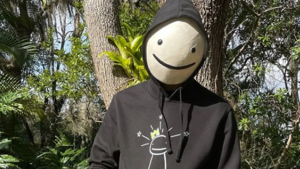 Person wearing a black hoodie with a sun graphic standing outdoors with a large round mask displaying a simple smiley face surrounded by greenery