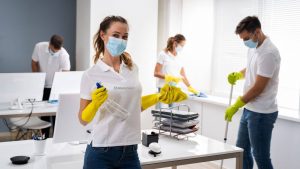 A team of office cleaners wearing white shirts and yellow gloves, cleaning an office space with desks, computers, and office supplies.