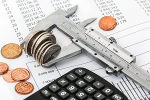 A close-up image of a metal vernier caliper measuring a stack of coins, alongside scattered coins, a calculator, and printed financial spreadsheets.