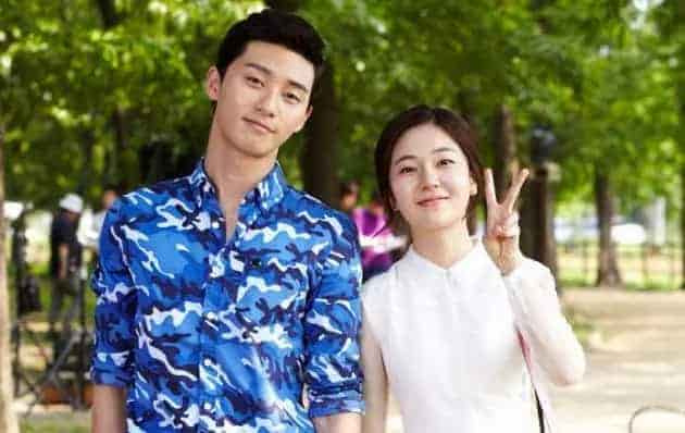 Park Seo Joon in a blue camouflage shirt alongside a smiling woman making a peace sign in a green park, hinting at a casual drama set or promotional event,
