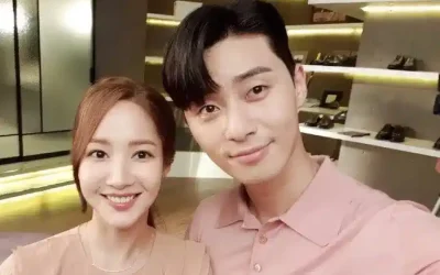 Park Seo Joon smiling with a woman in a pastel outfit during a 2025 brand event selfie, surrounded by shelves of shoes and bags, hinting at his endorsement career.
