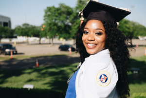 College Education - A person in a graduation cap and gown with a blurred background of trees and grass, facing away from the camera.