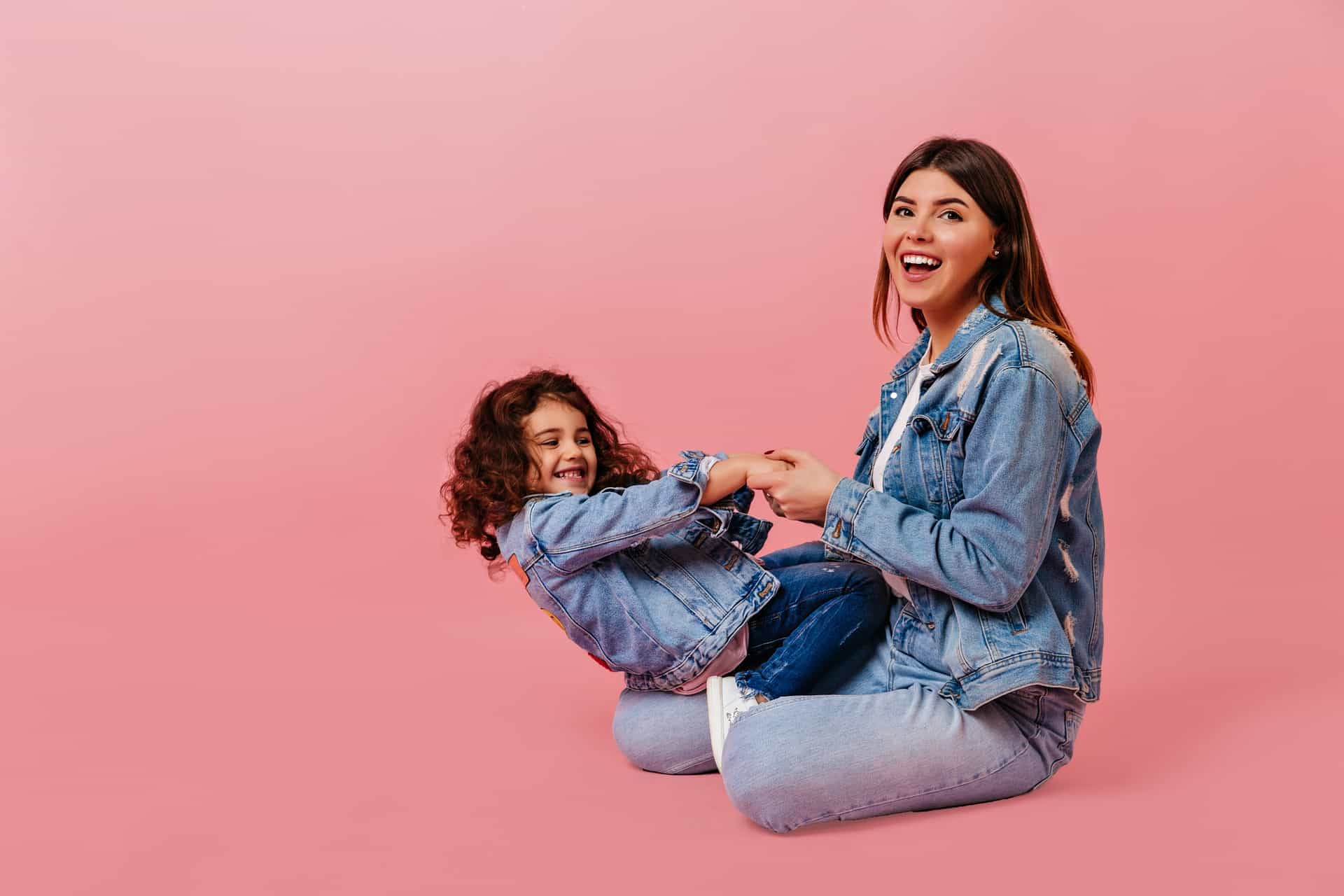Two individuals with obscured faces are sitting on a pink background, both wearing denim jackets and jeans. The adult is holding the child’s hands, and they appear to be engaged in an activity or interaction.