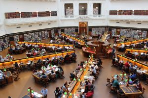 A spacious library reading room filled with numerous students seated at long wooden tables, studying and working on laptops, surrounded by bookshelves lined with books.