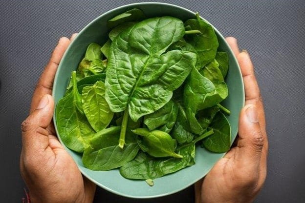 A pair of hands holding a light blue bowl filled with fresh spinach leaves against a dark grey background The spinach leaves are bright green and appear crisp indicating freshness