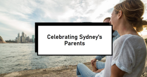 A person with their back facing the camera, looking towards a city skyline across a body of water, with a black rectangular overlay containing the text “Celebrating Sydney’s Parents.