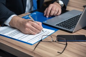 Close-up of a person’s hands writing on a form next to a laptop and eyeglasses on a desk.