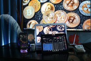 A collection of cryptocurrency coins with financial trading charts on smartphone and laptop screens, symbolising crypto trading.