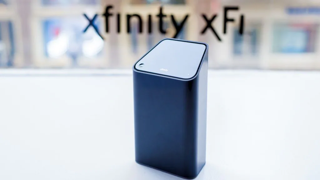The image depicts a black rectangular portable battery pack standing upright on a reflective surface. In the background, slightly out of focus, there’s a window with the words “xfinity xFi” visible, suggesting the setting may be a retail or service provider location. The battery pack has a smooth finish with rounded edges and features a prominent brand logo on its front side. ??