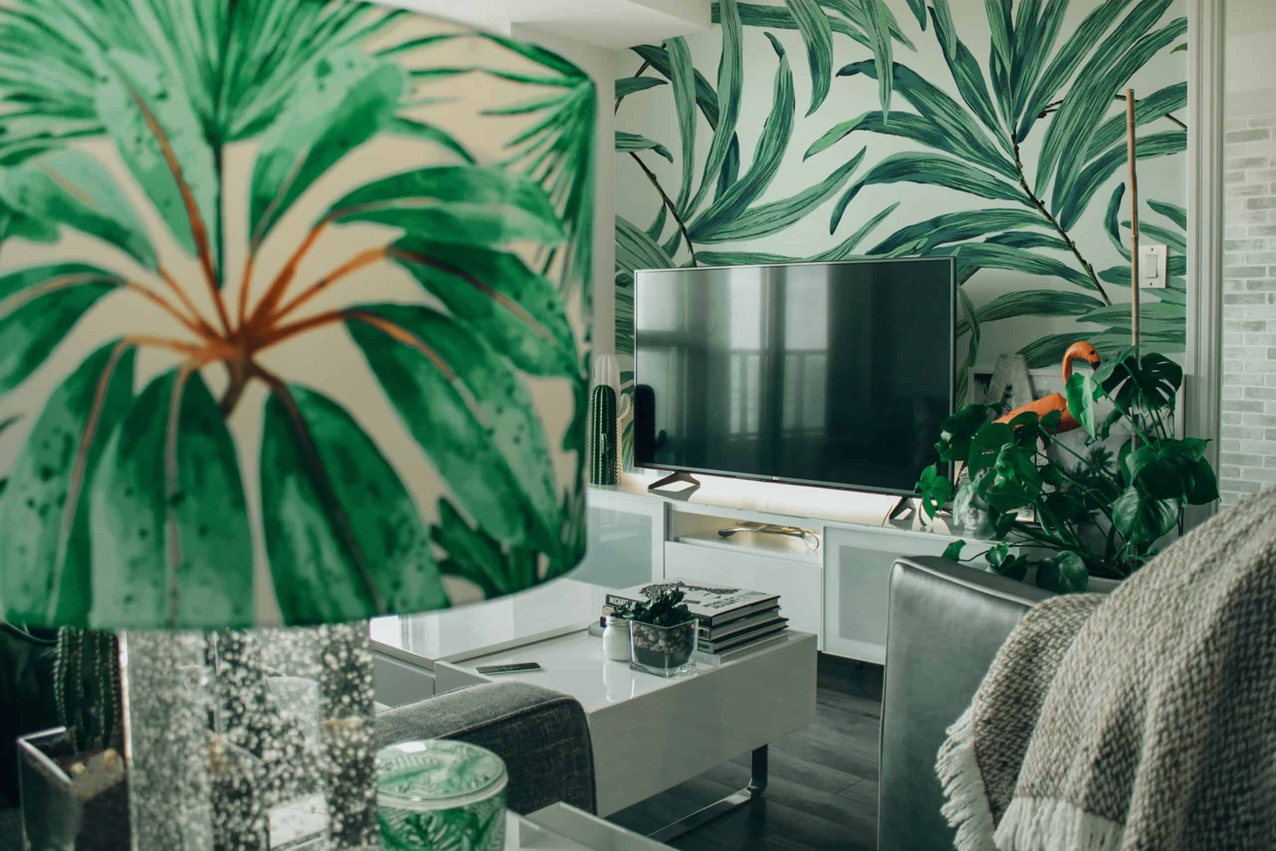 A stylish dining room featuring tropical leaf-patterned wallpaper and matching lampshade, with modern furniture and mounted TV.