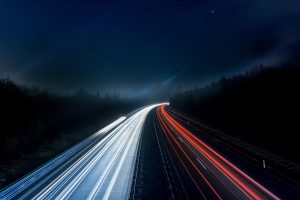 A long-exposure photograph capturing the bright white and red light trails of traffic on a highway at night, with a dark blue sky sprinkled with stars overhead.