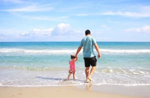 An adult and a young child holding hands and walking towards the sea on a sandy beach under a clear blue sky