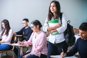 Students seated at desks in a classroom with one individual standing and holding study materials, representing an educational setting. #GAMSAT Tutor