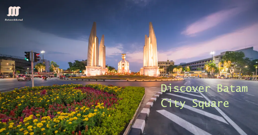 A panoramic view of Batam City Square at dusk, featuring a vibrant flowerbed in the foreground, a central fountain illuminated with warm lights, and two tall, pointed monuments symmetrically positioned on either side of the square. The sky is painted with hues of purple and blue during twilight. Text overlay on the image reads “Discover Batam City Square.