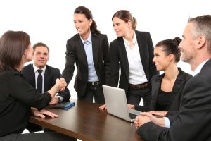 Business professionals engaged in a meeting, with one standing person shaking hands with another seated individual, indicating collaboration or agreement.