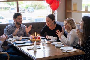 A group of people celebrating a birthday at a restaurant, gathered around a table with a cake topped with lit candles.
