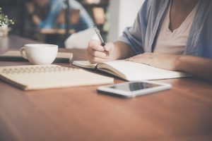 Person writing in a notebook with a smartphone and coffee cup on a wooden table.