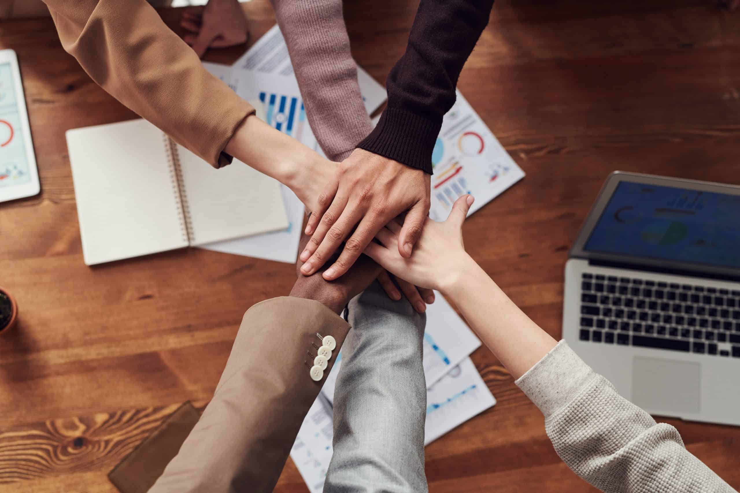 Team members joining hands over a worktable filled with business strategy documents and a laptop, symbolizing collaboration in SEO efforts.