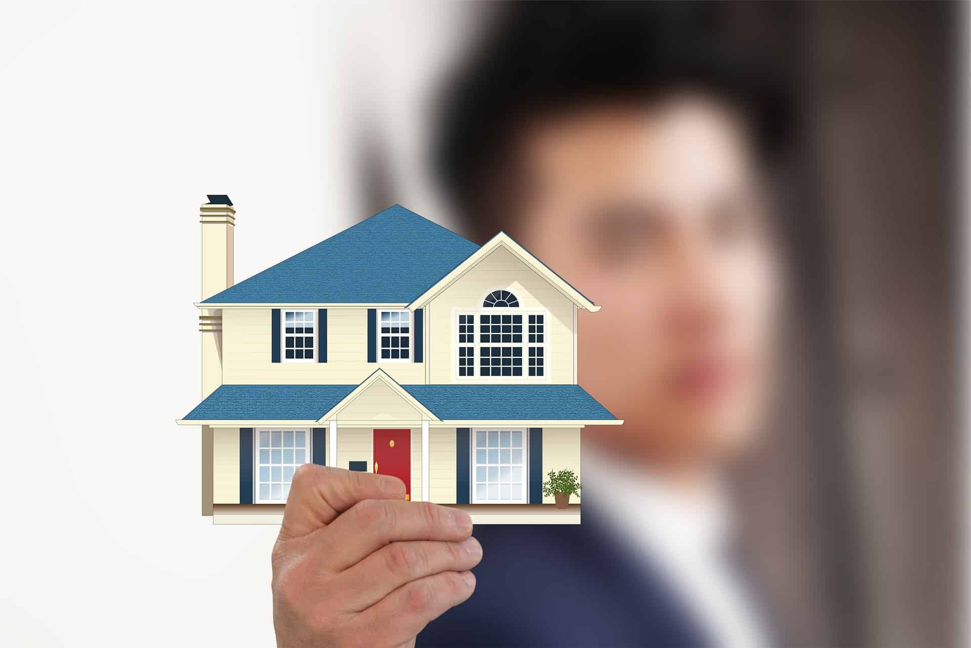 A hand holding a paper cut-out of a blue and white two-story house, symbolizing property investment.