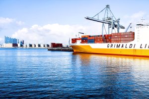 A large cargo ship from GRIMALDI LINES docked at a busy port loaded with colourful shipping containers under a clear blue sky. #Logistic Support