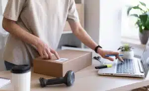 Person preparing a package for shipment with laptop and shipping tools on desk. #Dropshipping
