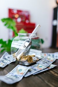 A glass jar filled with Bitcoin coins and water, surrounded by scattered U.S. hundred-dollar bills on a wooden surface.