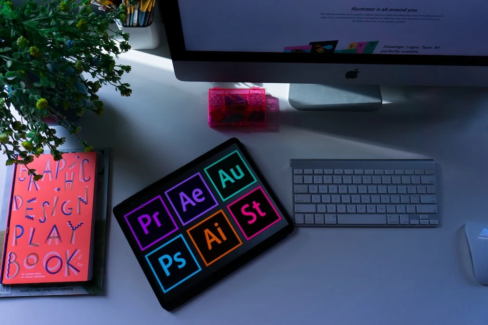 An illustration of a modern workspace with an illuminated keyboard featuring Adobe product shortcuts (Premiere Pro, After Effects, Audition, Photoshop, Illustrator, and Adobe Stock). The desktop computer displays ‘Illustrator for all around you,’ and a book titled ‘Graphic Design Play Book’ rests alongside a potted plant. This vibrant image highlights tools used by professionals in graphic design and multimedia industries.