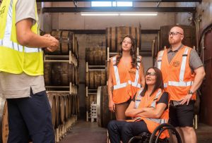 A warehouse scene with workers in orange safety vests, one of whom is in a wheelchair, highlighting accessibility and diversity in the industrial workplace.