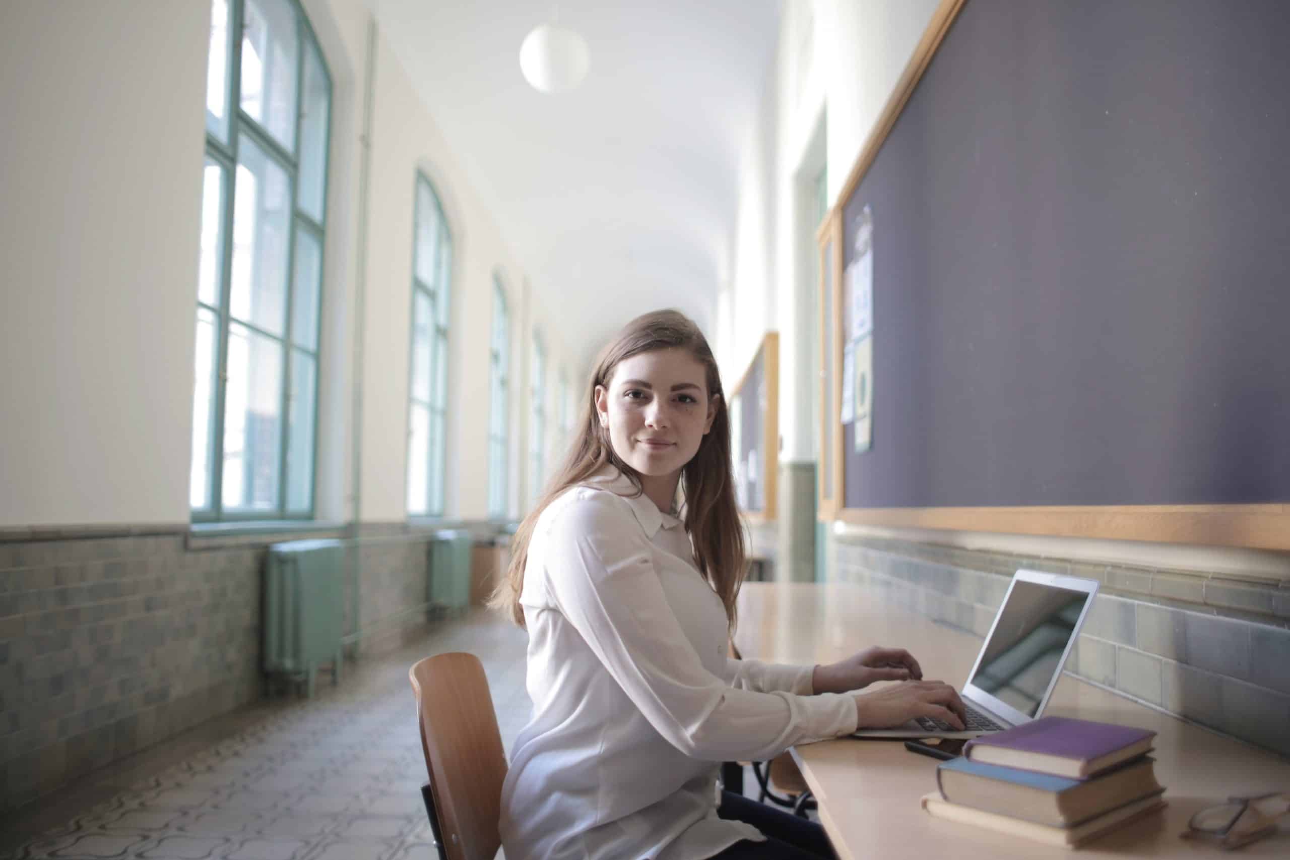 A person working on a laptop in a bright hallway with large windows, books stacked beside the laptop.