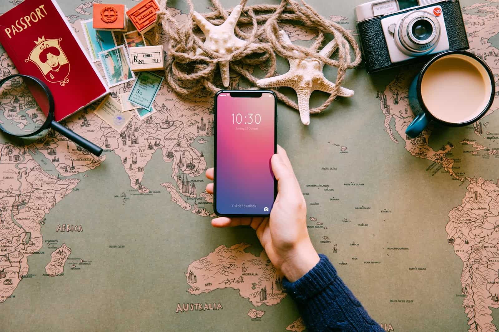 A traveler’s hand holding a smartphone over a world map surrounded by travel essentials like passport, camera, and currency.
