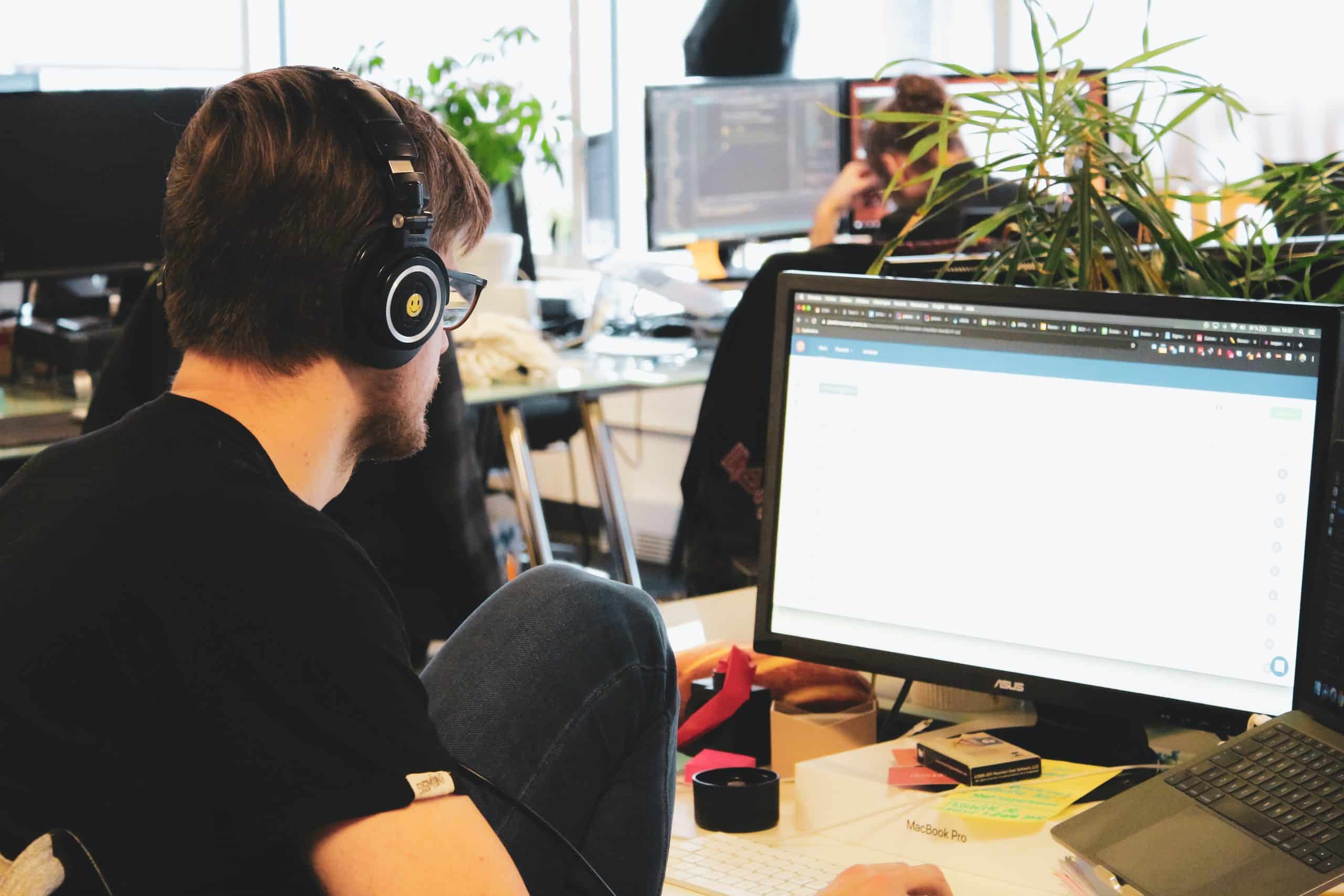 A person wearing headphones working on a computer in an office environment, with code visible on the screen in the background. #Combat Cyber Threats