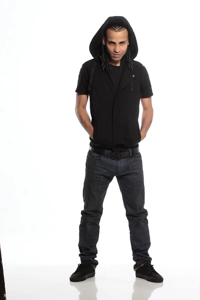 Person standing against a white background, wearing a black hooded top, black short-sleeved shirt, dark jeans, and black shoes, with hands in pockets and face obscured by pixelation.