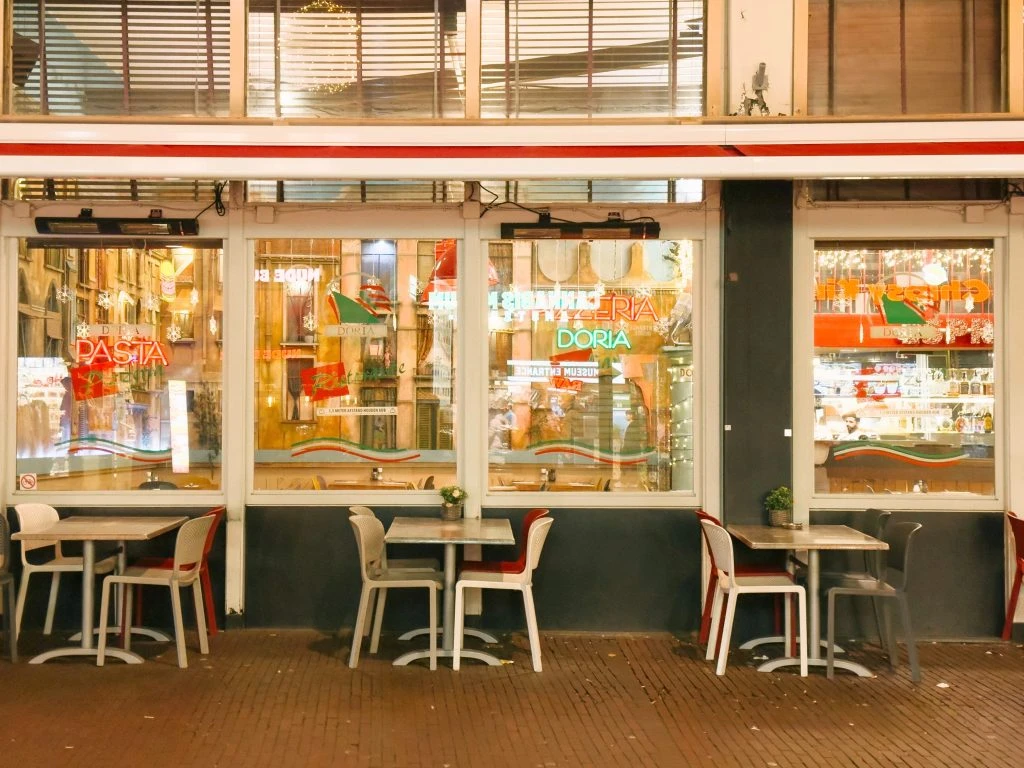 The exterior of a warmly lit restaurant named ‘PASTA DORIA’ at dusk, featuring large glass windows displaying shelves with bottles and other items. Outside, there are several white tables with chairs, suggesting an outdoor dining area.