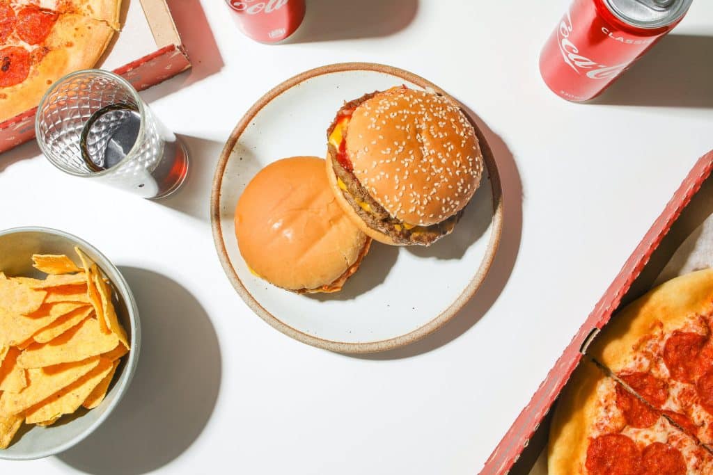 An overhead view of various fast food items including a cheeseburger with lettuce and sesame seed bun a plain hamburger a slice of pepperoni pizza a bowl of tortilla chips and two cans of Coca Cola on a white surface