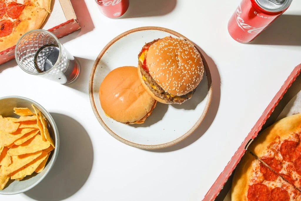An overhead view of various fast food items including a cheeseburger with lettuce and sesame seed bun, a plain hamburger, a slice of pepperoni pizza, a bowl of tortilla chips, and two cans of Coca-Cola on a white surface.