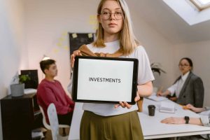 Person holding a tablet displaying the word "INVESTMENTS" in an office setting with colleagues in the background.