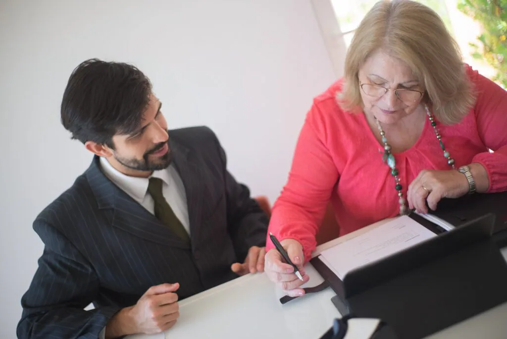 Two individuals with obscured faces are seated at a table, with one appearing to be signing or writing on a document. The person on the left is wearing a dark blazer and has dark hair, while the person on the right is wearing a red blouse and has blonde hair. A pen is in use by the individual in red, indicating an action of agreement or record-keeping. The setting suggests a formal or business-like interaction, possibly involving an agreement or transaction.