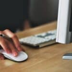 Close-up of a hand using a white computer mouse next to a keyboard and part of a monitor on a wooden desk. #Geraci Client Corner